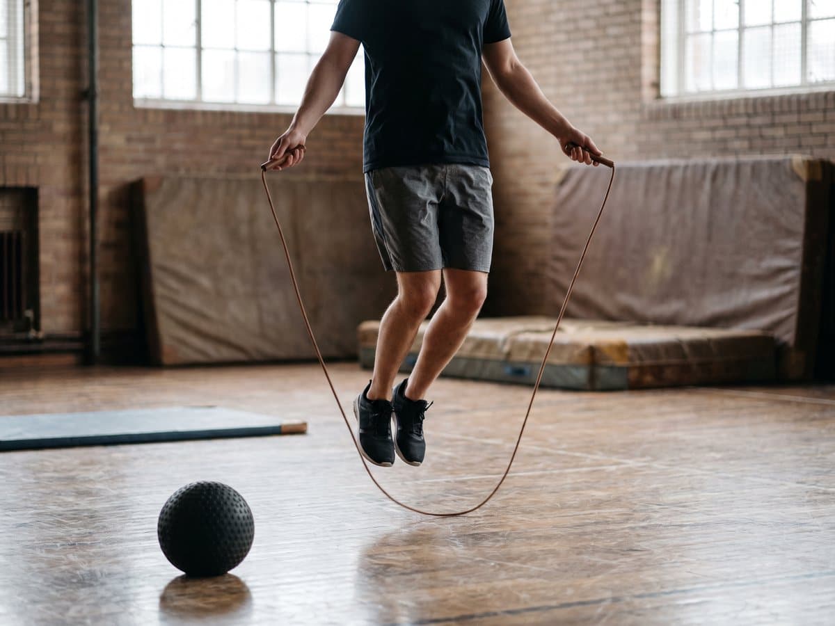A person mid-skip with a jump rope on a gym floor, a slam ball resting on the floor nearby