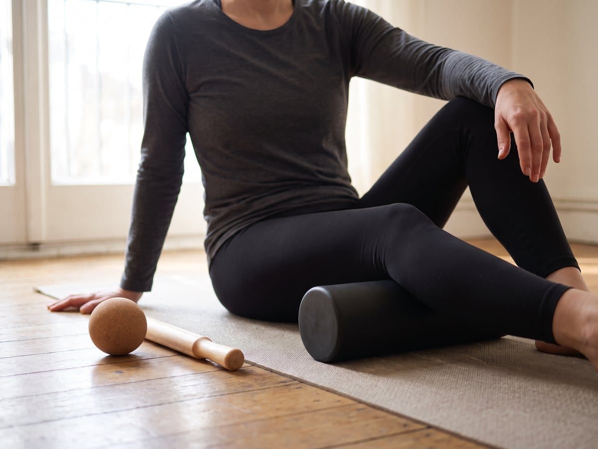 A person rolling a calf on a foam roller on an exercise mat, a massage ball and a roller stick on the floor beside them