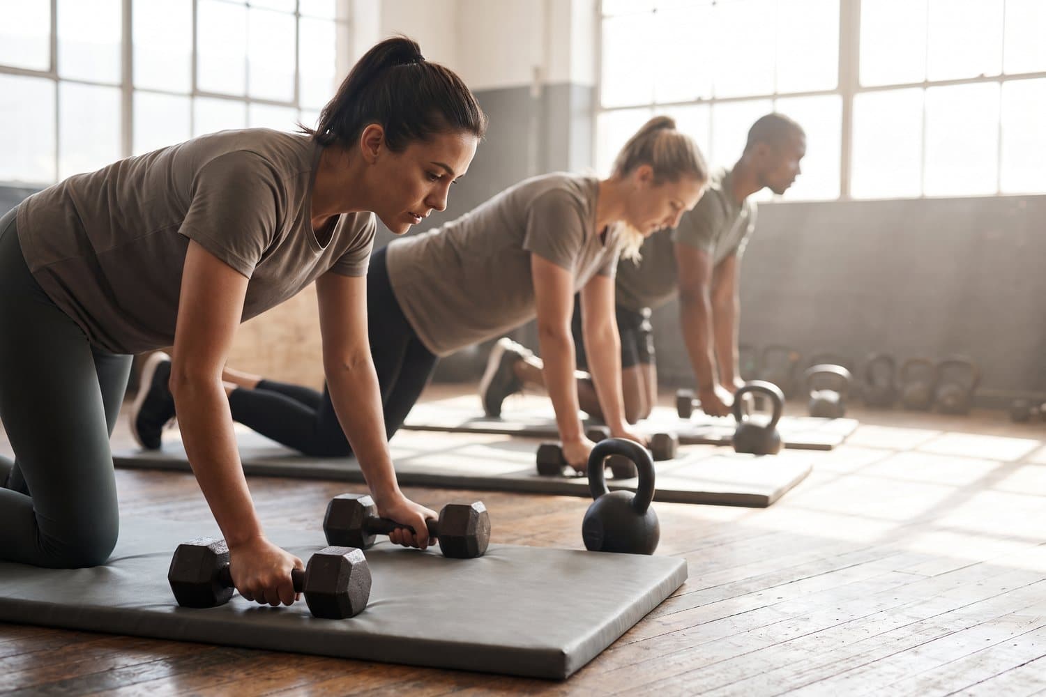 A few people training in a bright open gym with dumbbells, kettlebells and exercise mats, seen from the side